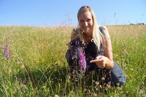 Eine blonde Frau kniet in einer blühenden Wiese und zeigt auf eine Blume