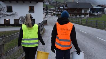 Zwei Menschen mit Warnwesten und Kübeln gehen die Straße runter