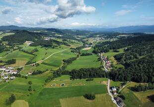 Luftbild das eine Mischung aus Wiesen, Waldstücken und Häusern entlang kleinerer Straßen